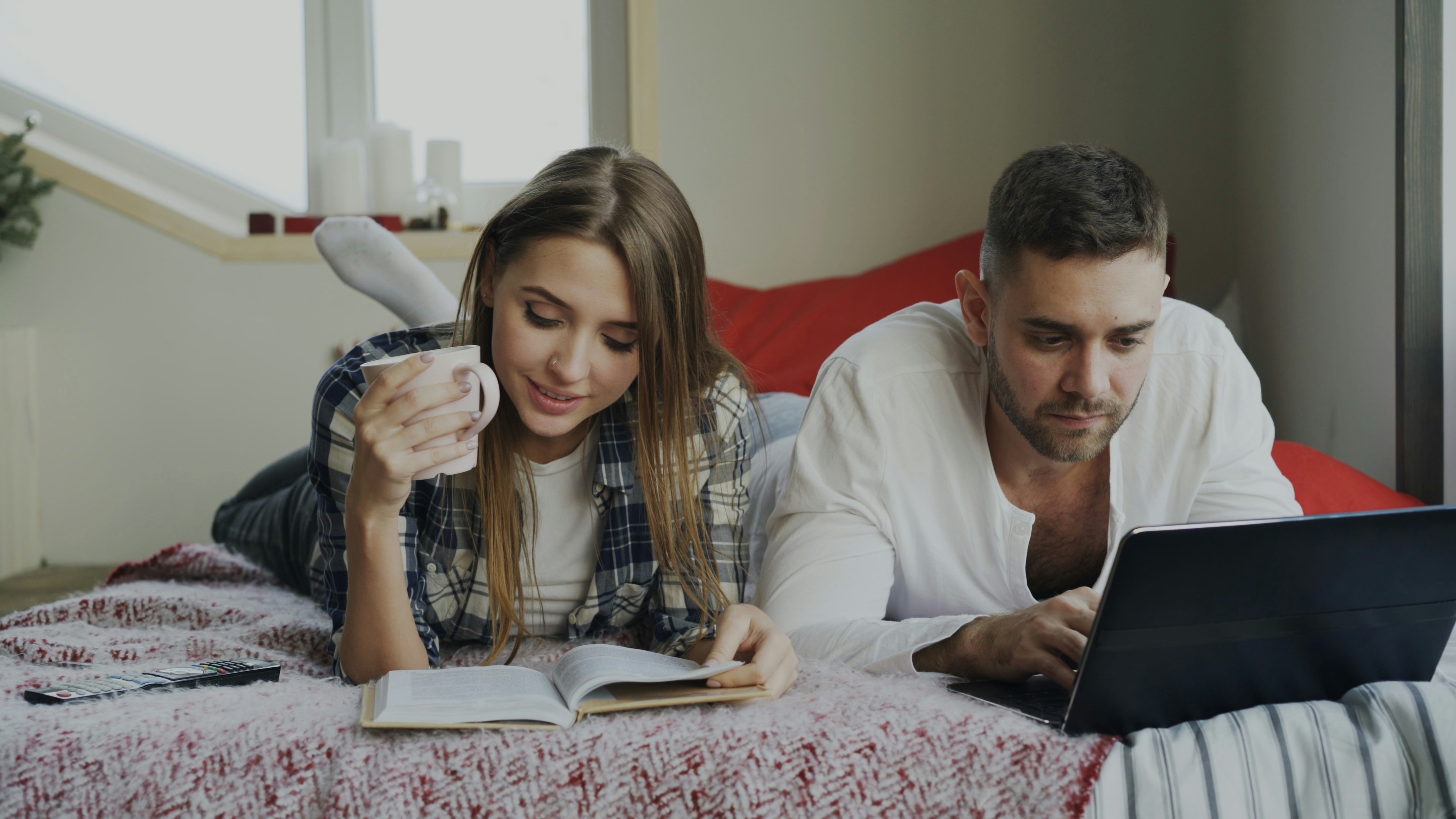 Cheerful couple have rest at home. Young man and woman lying in bed reading book and using laptop computer in the morning